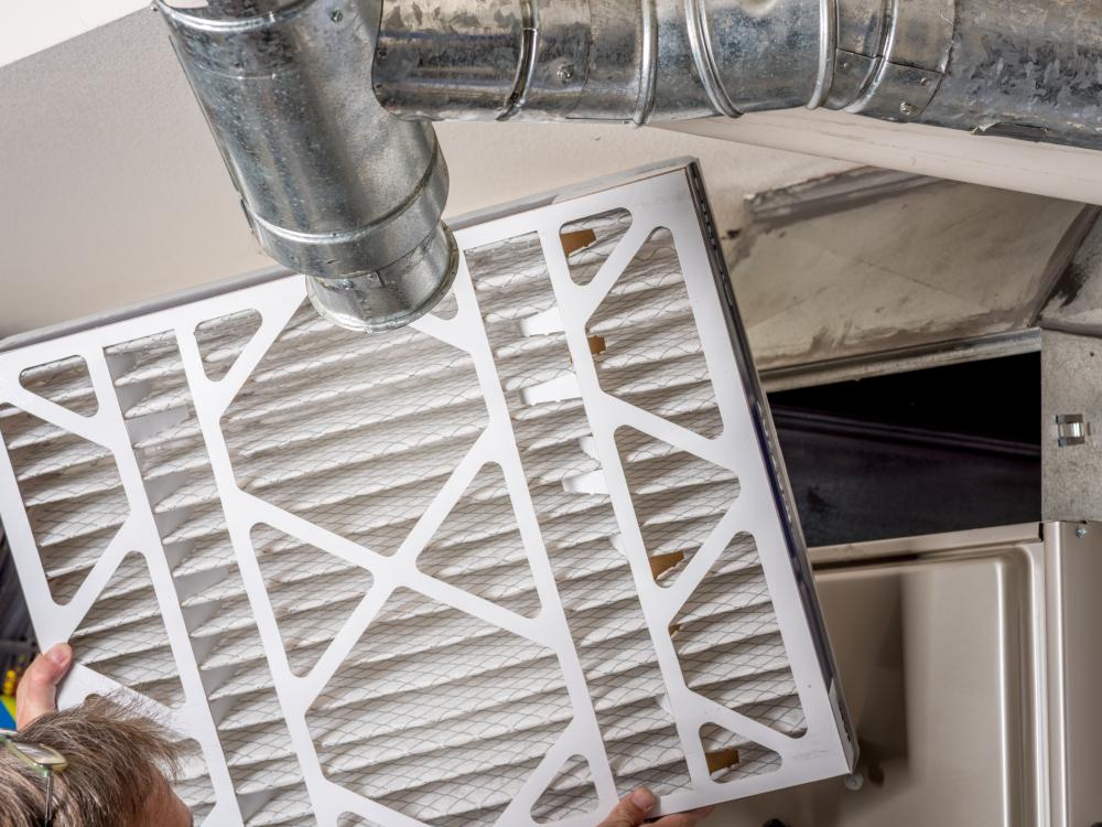 A person sliding a new air filter into a home HVAC furnace with metal ductwork overhead.