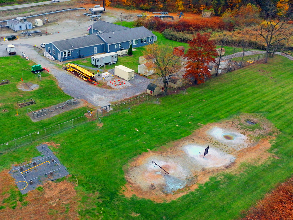 Aerial view of a facility with buildings, equipment, and fenced training or testing areas surrounded by grass and autumn trees.