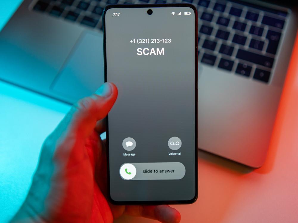 A hand holds a smartphone displaying an incoming call labeled “SCAM” in front of a laptop.