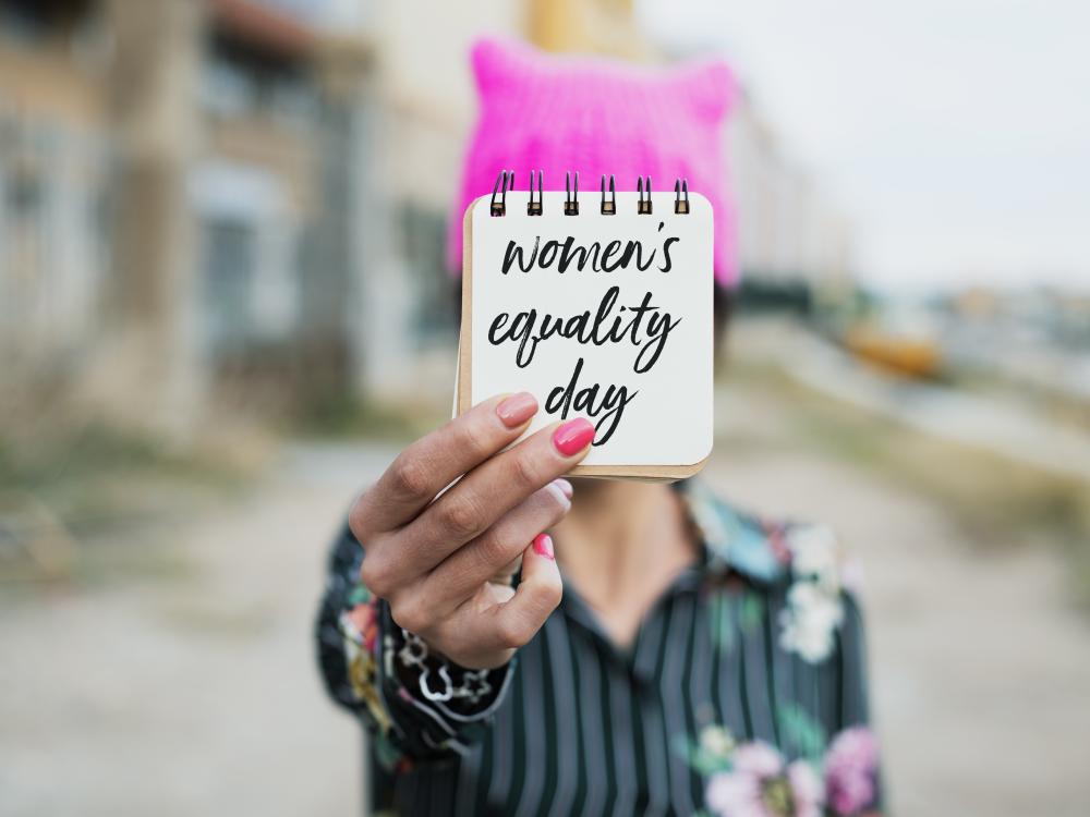 Person holding a small notebook in front of their face with the words “women’s equality day” written on it.