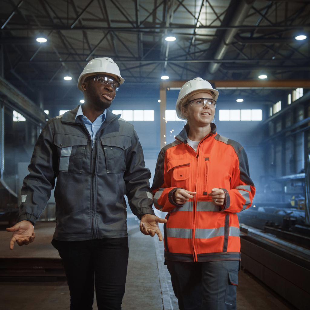 Two workers wearing hard hats and safety glasses walk and talk inside an industrial facility, surrounded by metal beams and machinery.