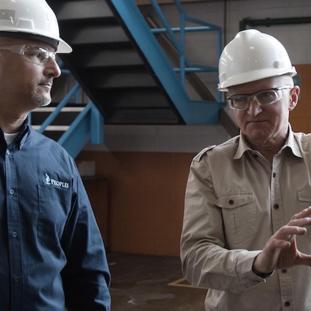 Two workers wearing hard hats and safety glasses stand inside an industrial facility, discussing equipment while one gestures toward machinery.