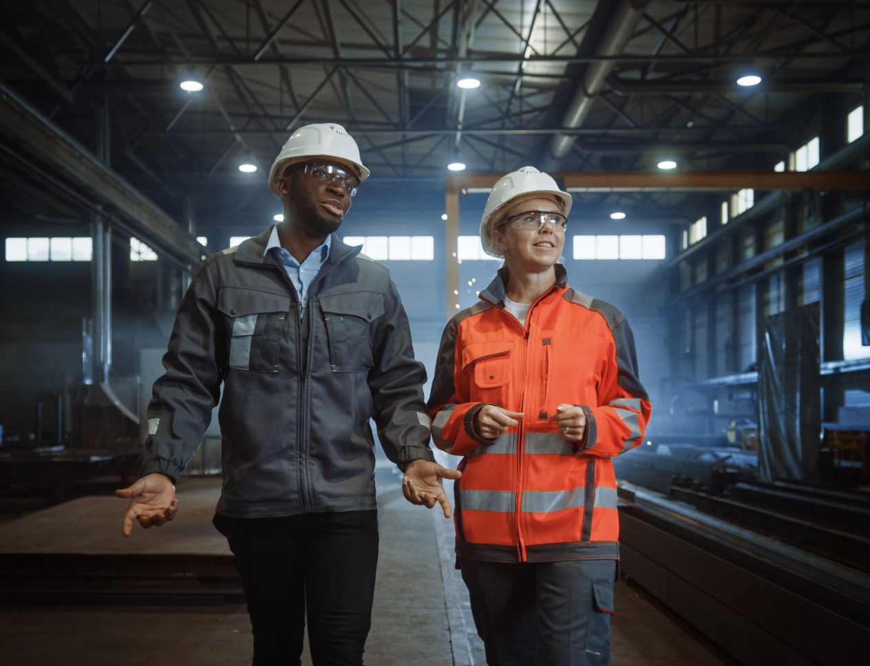 Two workers wearing hard hats and safety glasses walk and talk inside an industrial facility, surrounded by metal beams and machinery.