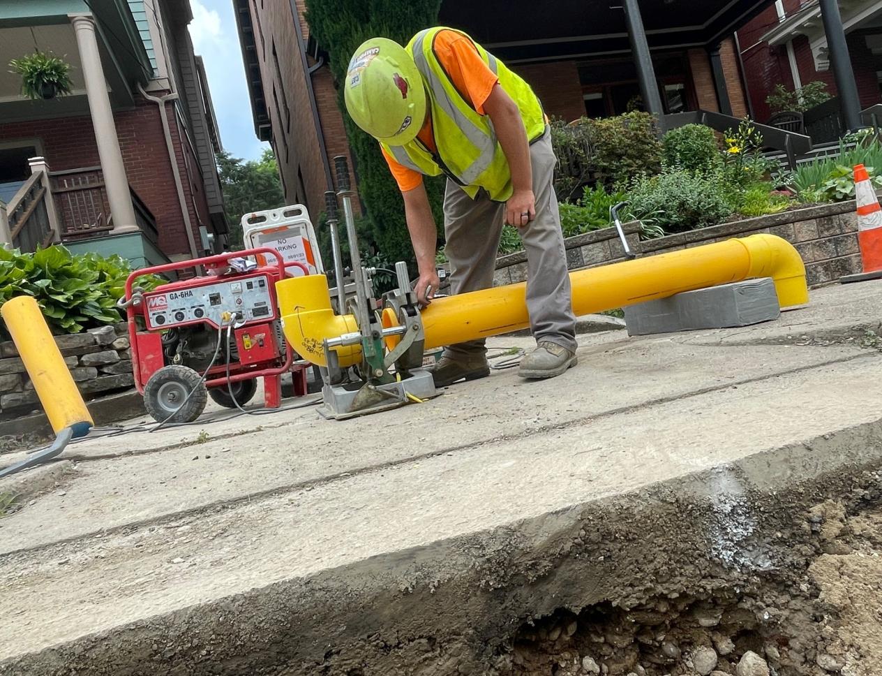 A construction worker in a yellow vest and hard hat operates equipment on a sidewalk, near a bright yellow pipe.