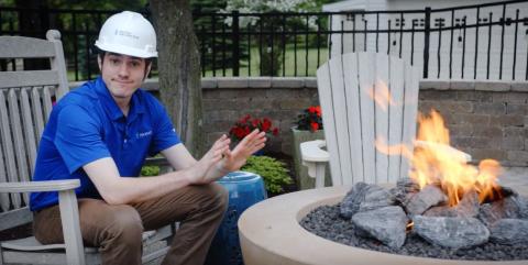 Peoples Natural Gas employee wearing a hard hat sits beside an outdoor fire pit, warming his hands near the flame in a backyard setting