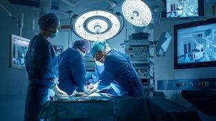 Surgical team in an operating room performing a procedure under bright overhead lights, surrounded by medical monitors and equipment.